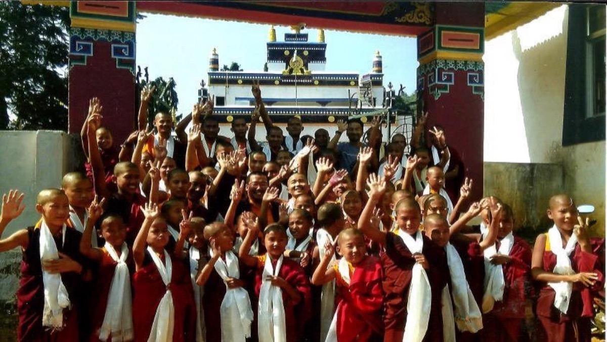Young monks in maroon robes raising their hands joyfully at the monastery gate